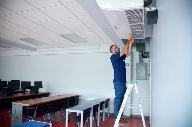 Un funcionario del Campus Tecnológico Local San José está subido en una escalera dentro de un laboratorio de computación dándole mantenimiento a un aire acondicionado