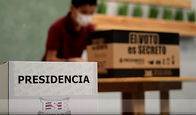 Una urna electoral en primer plano y de fondo la silueta difuminada de un niño emitiendo su voto.