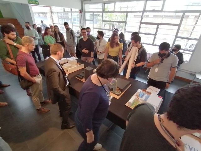 Mesa alargada en el primer piso del edificio de la Escuela de Arquitectura y Urbanismo con siete de las catorce maquetas presentadas. Se ve mucha gente alrededor de ellas.