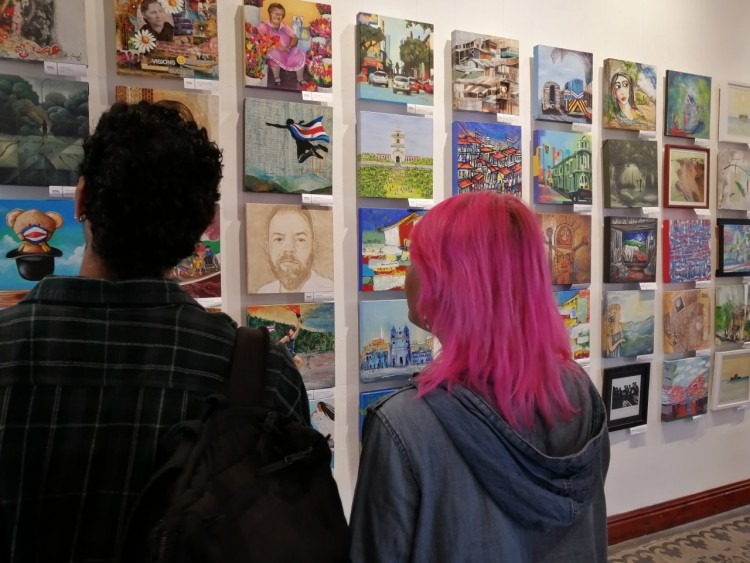 Dos personas observan una pared llena de pinturas en diferentes técnicas.
