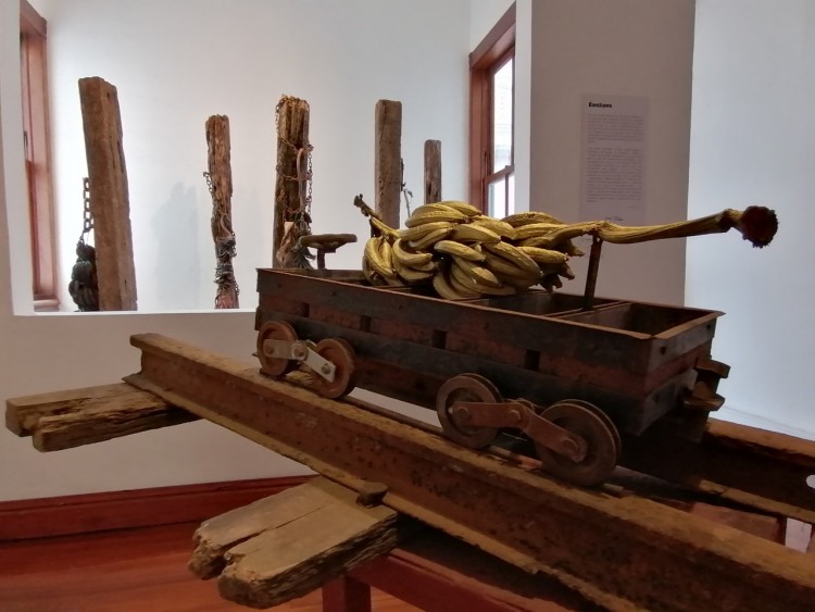 Una exposición artística donde se aprecia un vagón en miniatura de ferrocarril sobre dos rieles y cargado de banano.