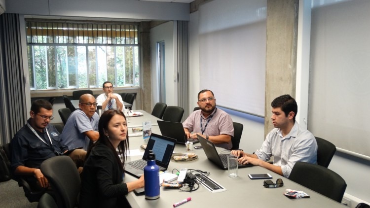 Cinco hombres y una mujer observando una presentación en una mesa de sesiones. 