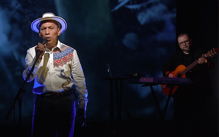 El cantante frente al micrófono, con sombrero. Al fondo, lo acompaña un músico con guitarra.