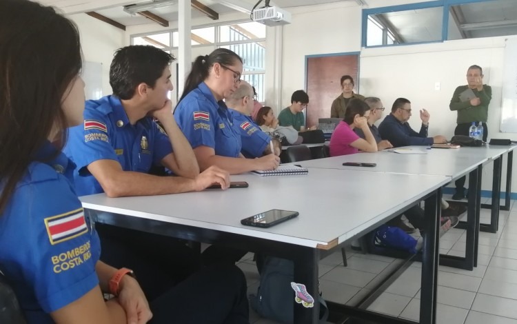 Un aula de Arquitectura y Urbanismo del TEC donde hay cuatro funcionarios del Cuerpo de Bomberos sentados en primera fila tomando apuntes de una exposición de los docentes. Atrás se ven algunos estudiantes.