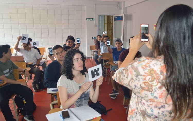 Imagen de una mujer que recoge la información con su celular y sus alumnos  están sentados con la respuesta en una hoja mostrándola hacia el frente.