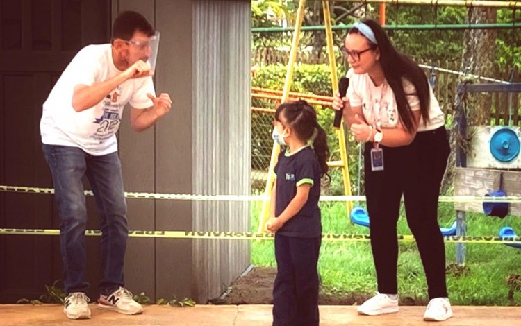 Niña aprende señas básicas con el invitado. Están en la plazoleta del kínder. 