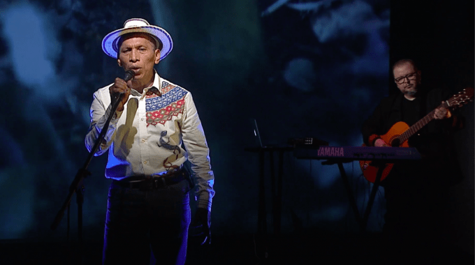 El cantante frente al micrófono, con sombrero. Al fondo, lo acompaña un músico con guitarra.