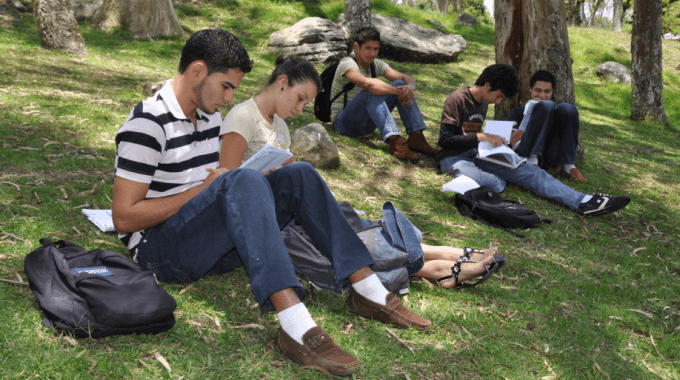 Cinco estudiantes del TEC se encuentran (sentados) estudiando, en la zona verde de esta Institución  Son cuatro  hombres y una mujer.