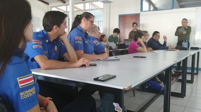 Un aula de Arquitectura y Urbanismo del TEC donde hay cuatro funcionarios del Cuerpo de Bomberos sentados en primera fila tomando apuntes de una exposición de los docentes. Atrás se ven algunos estudiantes.