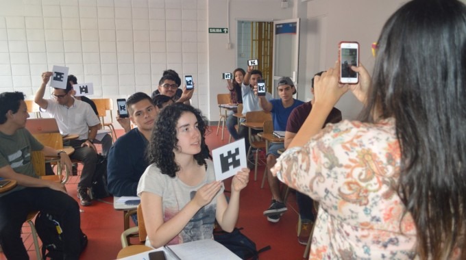 Imagen de una mujer que recoge la información con su celular y sus alumnos  están sentados con la respuesta en una hoja mostrándola hacia el frente.