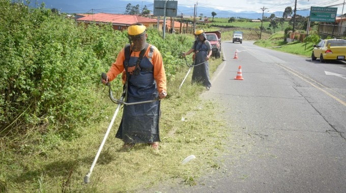 Dos personas realizan corte de zacate al lado del camino.