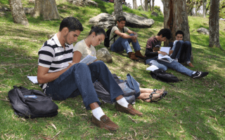 Cinco estudiantes del TEC se encuentran (sentados) estudiando, en la zona verde de esta Institución  Son cuatro  hombres y una mujer.