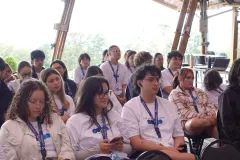 Grupo de estudiantes sentados con camisetas blancas del evento &amp;quot;Unnatural Selection, Synbiothon&amp;quot;, con gafetes azules, atentos a una presentación.