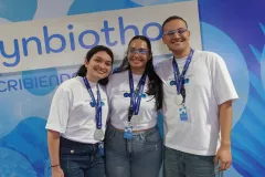 Tres jóvenes con camisetas blancas y medallas de plata, posando frente al backdrop de Synbiothon 2026.