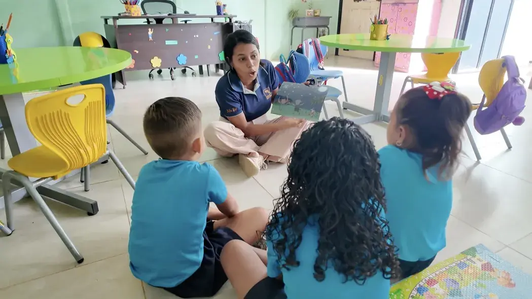 Maestra leyendo un cuento ilustrado a tres niños sentados en el suelo del aula.